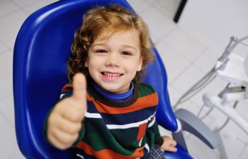 A smiling little boy in a dental chair showing his thumb up.