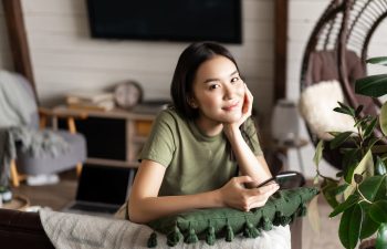 A young woman in a green t-shirt sits indoors, leaning on a cushion, holding a smartphone, and looking at the camera. A laptop and home decor are visible in the background., 