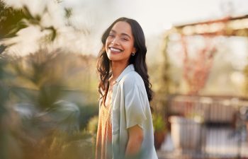 A woman with long dark hair smiles while standing outdoors on a sunny day, surrounded by greenery and blurred plants, with soft sunlight in the background.