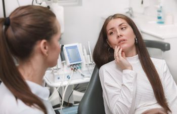 A woman in a dental chair touches her cheek while talking to a dentist in a clinic setting., 