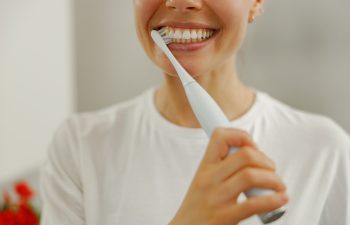A person wearing a white shirt is smiling while brushing their teeth with an electric toothbrush. Only the lower half of their face is visible.